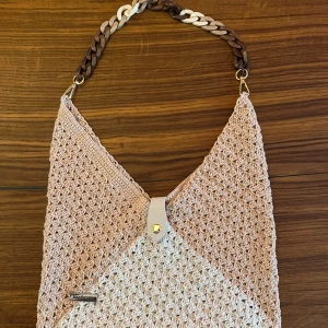 Beige stickad axelväska med kedja - Snygg stickad axelväska i beige och ljusrosa toner med grovt mönster. Väskan har en cool flätad kedja i brunt, beige och vitt som axelrem och en liten läderdetalj med knapp framtill. Perfekt för dig som gillar unik design och handgjorda accessoarer.