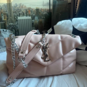 Ljusrosa quiltad YSL axelväska - Säljer en ljusrosa quiltad axelväska från YSL med silverfärgad kedja och ikonisk YSL-logga framtill. Väskan är tillverkad i mjukt skinn och har magnetlås samt flera fack inuti. Perfekt storlek för det viktigaste och ger en lyxig vibe till din outfit.