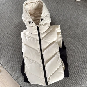 Beige dunväst med huva från Zara - Säljer en beige dunväst från Zara i storlek XS. Västen har en stor, mysig huva med snörning och svart dragkedja framtill. Sidorna är svarta vilket ger en snygg kontrast. Perfekt för lager-på-lager och chill stil under höst och vinter.Helt oanvänd