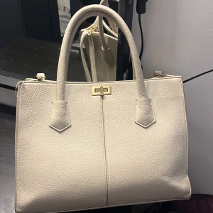 Beige handväska med gulddetaljer - Säljer denna fina beige handväska i skinnimitation med guldfärgat lås och detaljer. Väskan har både handtag och axelrem, samt ett rymligt fack med dragkedja på insidan. Perfekt väska för skolan och väskan är i bra skick!