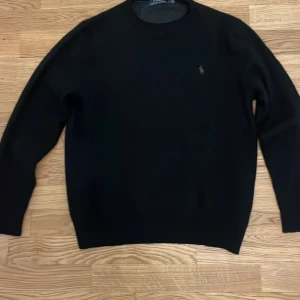 Svart stickad tröja från Polo Ralph Lauren - Svart stickad tröja från Polo Ralph Lauren med klassisk rund hals och diskret broderad logga på bröstet. Tröjan har långärmade och en clean, enkel design som funkar till allt. Perfekt för dig som gillar stilrena plagg med premiumkänsla.