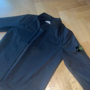overshirt  Stone Island S - Svart overshirt från Stone Island med klassisk krage, bröstficka och ikonisk patch på vänster ärm. Jackan har dragkedja framtill och är tillverkad i ett slätt syntetmaterial. Passar runt 180 cm Skickas med tag och sin påse, skriv vid någon fundering🙌