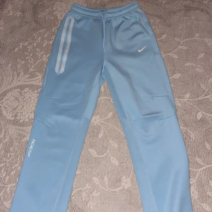 Nike NOCTA ljusblå mjukisbyxor - Nike NOCTA mjukisbyxor i ljusblå färg med vit Swoosh-logga på höften och NOCTA-tryck på benet. Byxorna har elastisk midja med snörning och raka ben. Tillverkade i mjukt syntetmaterial som är skönt mot huden. Perfekt för chill eller träning.