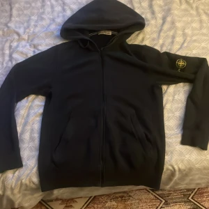 Svart hoodie från Stone Island - Svart hoodie från Stone Island med klassisk kompasspatch på vänster ärm. Tröjan har huva, dragkedja framtill och två fickor. Perfekt för dig som gillar streetwear och vill ha något stilrent med ikonisk logga. Materialet är mjuk bomull som känns skönt mot huden. Pris kan diskuteras 