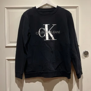 Svart sweatshirt från Calvin Klein Jeans - Svart sweatshirt från Calvin Klein Jeans i storlek S. Tröjan har ett stort CK-logotryck i vitt och silver på bröstet. Rund halsringning och långa ärmar med ribbade muddar. Perfekt för en chill och stilren look.