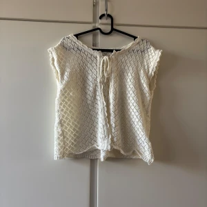 Vit stickad blus med knyt - Supersöt vit stickad blus med korta ärmar och knyt framtill. Blusen har ett luftigt, virkat mönster och vågig kant nertill och vid ärmarna. Perfekt för varma dagar och ger en romantisk vibe till din outfit.