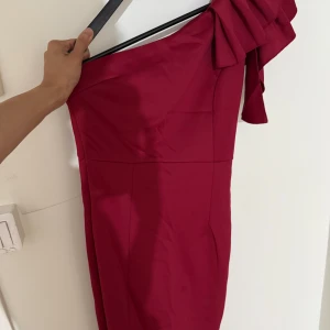 Vinröd oneshoulder långklänning  - Säljer en vinröd kortklänning med oneshoulder-design och en snygg volangdetalj på axeln. Klänningen har en figurnära passform och är perfekt för dig som vill sticka ut med en cool och elegant look. Materialet känns mjukt och har en lätt glans.