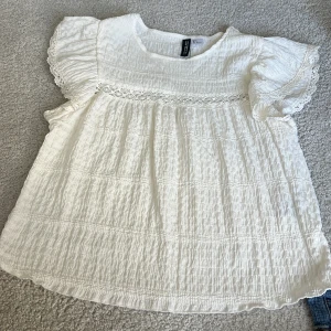 Vit blus med spetsdetaljer H&M - Superfin vit blus från H&M med korta volangärmar och spetsdetaljer vid axlar och bröst. Blusen är gjord i ett krinklat bomullstyg som ger en luftig känsla. Perfekt till jeans eller kjol för en söt och avslappnad look.