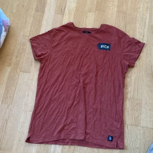 Röd t-shirt från Saligia med tryck - Säljer en röd t-shirt från Saligia med coolt tryck på bröstet och ryggen. Framsidan har en liten patch med texten 'RICH' och baksidan har ett stort statement-tryck med texten 'LIVING LARGE SPENDING TIME NOT MONEY' och en illustration av ett timglas med vingar. Skön bomullskvalitet och klassisk passform.