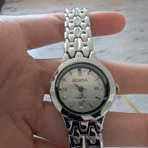 Silverfärgad ROSRA damklocka - Stilren damklocka från ROSRA med rund urtavla och romerska siffror. Klockan har ett coolt armband i silverfärgad metall med snyggt mönstrade länkar och klassiskt spänne. Urtavlan är ljus med diskreta detaljer och quartz-urverk. Kan ta av länkarna men vet själv inte hur man gör det. Aldrig använd och inga defekter