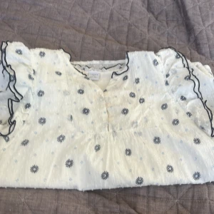 Vit blommig blus med volang - Supersöt vit kortärmadblus från Lindex med marinblå och ljusblå blommor. Blusen har volangdetaljer på ärmarna och runt halsen, samt en lätt v-ringning. Materialet är tunt och luftigt, perfekt för varma dagar. Passformen är normal och ärmarna är korta med snygga volanger.