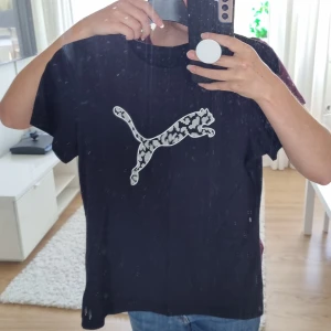 Svart tshirt från Puma  - Svart t-shirt från Puma i bomull med klassisk rund hals och korta ärmar. På bröstet finns en stor Puma-logga med leopardmönster i vitt och svart, vilket ger en cool och sportig vibe. Passformen är normal och materialet känns mjukt mot huden 💓 Skriv till mig vid köp eller fråga! 