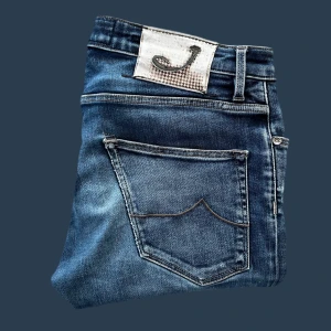 Jacob Cohën blå jeans straight fit - Snygga blå jeans från Jacob Cohën med klassisk femficksdesign och diskreta slitningar. Jeansen har raka ben och normal passform, med detaljer som kontrastsömmar och logotyp på bakfickan. Tillverkade i mjuk bomullsdenim för en bekväm känsla. Liten slitning på knät som syns i bilden