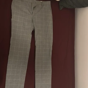 Grå rutiga kostymbyxor - Snygga grå kostymbyxor med vitt rutmönster och klassisk rak passform. Byxorna har bälteshällor, diskret dragkedja och är perfekta för en clean och stilren look. Materialet känns mjukt och lätt, vilket gör dem sköna att bära hela dagen.