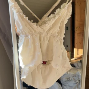 Vit linnetopp med volang och broderi - Supersöt vit linnetopp med tunna axelband som korsas i ryggen. Toppen har volangkant vid halsen och små broderade detaljer över hela tyget. Den är lätt och luftig med en romantisk vibe, perfekt för varma dagar.