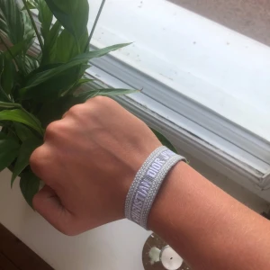 Grått armband från Christian Dior - Säljer ett snyggt grått armband från Christian Dior med texten 'CHRISTIAN DIOR J'ADIOR' broderad i vitt. Armbandet är gjort i tyg och har en tofsdetalj i ena änden. Perfekt accessoar för att lyfta din outfit. Skriv privat vid frågor och funderingar! Pris går att diskuteras.