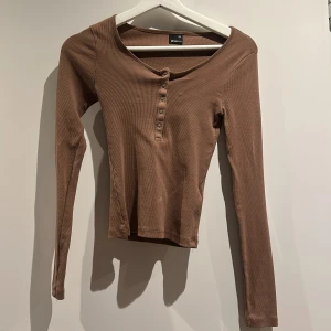 Brun ribbad långärmad topp Gina Tricot - Säljer en brun långärmad topp från Gina Tricot i storlek XXS. Toppen är ribbad med rundad hals och har knappar framtill som snygg detalj. Materialet är mjukt och stretchigt,