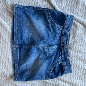 Blå jeanskjol med fickor - Säljer en klassisk blå jeanskjol med snygga fram- och bakfickor samt knappstängning framtill. Kjolen har en rak och kort passform, perfekt för dig som gillar en enkel och cool stil. Passar till sneakers eller boots för en avslappnad vibe.