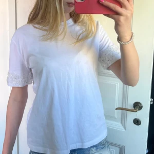 Vit t-shirt med pärlor från Zara - Vit t-shirt från Zara i mjuk bomull med korta ärmar. Ärmarna har detaljer med pärlor, blommor och paljetter som ger en extra touch. Perfekt för dig som gillar att sticka ut med snygga detaljer. Rund hals och normal passform.