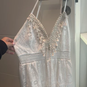 Vit spetsklänning med snörning och pärlor - Superfin vit långklänning i spets med bohemisk känsla. Klänningen har smala axelband, v-ringad omlott med flätad snörning och tofsar dekorerade med pärlor och snäckor. Spetsen har blommigt mönster och detaljer i guldtråd. Perfekt för sommarens alla tillfällen!