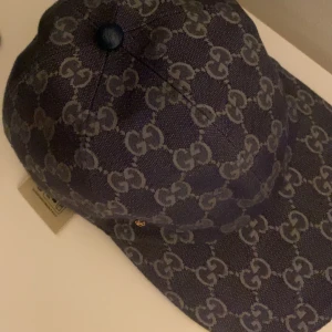 Blå Gucci keps med GG-mönster - Snygg blå keps från Gucci med det klassiska GG-mönstret över hela kepsen. Justerbar rem baktill i mörkblått och insida i beige bomullstyg. Perfekt för dig som vill ha en lyxig och trendig accessoar med tydlig designerstil.