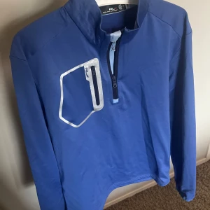 Blå Ralph Lauren tröja med half zip (golf) - Snygg blå långärmad tröja från RLX Ralph Lauren med half zip framtill och coolt vitt RLX-tryck på bröstet. Tröjan har en sportig look med praktisk bröstficka och är gjord i mjukt polyester-material. Perfekt för dig som gillar sport, speciellt golf.