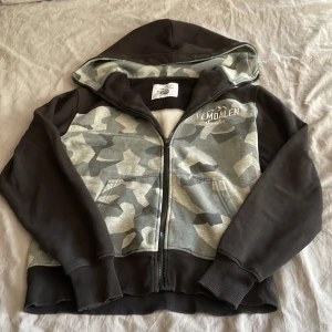 Camouflage hoodie från United by Nature - Säljer en hoodie från United by Nature med Vemdalen Sweden-brodyr. Tröjan har grått och svart camouflage-mönster, svart huva och ärmar, samt dragkedja framtill. Mjuk insida och ribbade muddar. Perfekt för chill dagar och outdoor vibes. Storlek S