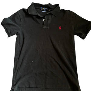 Svart Polo Ralph Lauren t-shirt  - Klassisk svart t-shirt från Polo Ralph Lauren i bomull med korta ärmar och krage. Röd broderad logga på bröstet ger en clean och stilren vibe. Classic fit som sitter snyggt utan att vara för tight. Perfekt till jeans eller shorts.