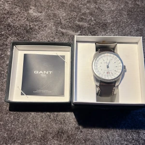 GANT herrklocka med brun läderrem - Stilren herrklocka från GANT med vit urtavla och detaljer i silver. Klockan har en rund boett i metall och en mörkbrun läderrem med ljusa sömmar. Urtavlan visar flera tidszoner och har datumvisning. Perfekt accessoar för dig som gillar klassisk och tidlös stil. Batteribyte behövs göras!