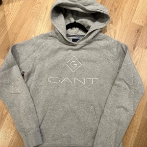 Grå hoodie från GANT med logga - Snygg grå hoodie från GANT med stor vit logga på bröstet. Tröjan har huva, magficka och långa ärmar. Mjuk och skön bomullsmix som passar perfekt när du vill vara chill. Klassisk och enkel design som funkar till jeans eller mjukis.