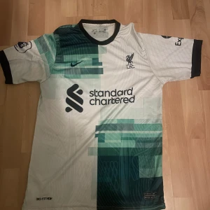 Liverpool Salah bortatröja Nike L - Liverpool FC bortatröja med M. Salah 11 på ryggen. Designad av Nike i Dri-FIT-material, vit bas med gröna och mörkblå grafiska block, svarta detaljer och klubbmärke broderat på bröstet. Kortärmad med svart krage och muddar. Snygg och modern look. Storleken är liten och passar både storlek M och S