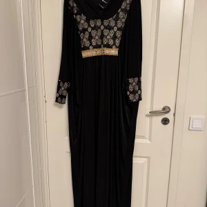 Svart långklänning med gulddetaljer  - Svart långklänning med glittriga gulddetaljer och silvermönster på bröstet och ärmsluten. Klänningen har en bred, draperad halsringning och långärmade ärmar. Materialet är mjukt och faller snyggt, perfekt för dig som gillar eleganta och unika plagg. Oanvänd.