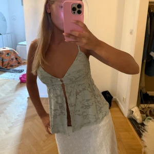 Ljusgrön spetsig linnetopp med knäppning - Supersöt ljusgrön linnetopp med tunna axelband och v-ringning. Toppen har spetsdetaljer och en liten knapp framtill som ger en öppen effekt. Fladdrig och luftig passform, perfekt för varma dagar.