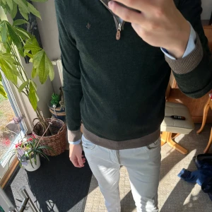 Mörkgrön half zip tröja från Morris - Snygg mörkgrön tröja från Morris med half zip och brunt ribbad kant vid ärmslut och nederkant. Liten Morris-logga broderad på bröstet. Perfekt att bära över skjorta för en chill och stilren look. Långärmad och i merinoull som känns skönt mot huden. Knappt använd.