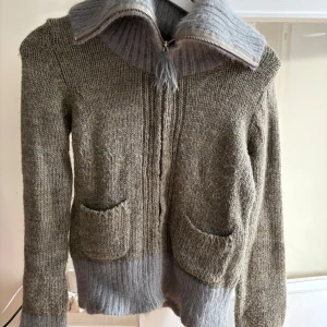 Grå stickad kofta Odd Molly - Säljer en mysig grågrön stickad kofta från Odd Molly med bred ribbad krage och dragkedja framtill. Koftan har två fickor och kontrastfärgade muddar i ljusgrått. Materialmix med ull, alpacka och akryl, perfekt för kyliga dagar.