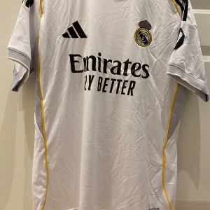 Real Madrid vit matchtröja Adidas - Säljer en officiell Real Madrid fotbollströja från Adidas med svart logga, klubbmärke och sponsortryck. Tröjan är vit med gula detaljer längs sidorna och har korta ärmar med ribbade muddar. Materialet är lätt och andas, Aeroready-teknologi för extra komfort.