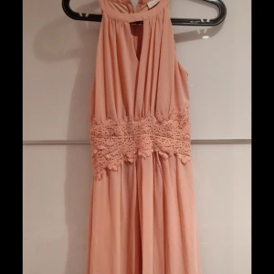 Ljusrosa långklänning med spetsdetalj - Säljer en elegant ljusrosa långklänning med vacker spetsdetalj i midjan. Klänningen har ärmlös design och ett mjukt, flytande tyg som ger en snygg siluett. Perfekt för dig som vill ha en romantisk och feminin look med extra detaljer.