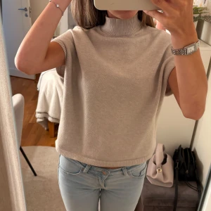 Beige stickad polotröja med kort ärm - Säljer en beige stickad polotröja med korta ärmar och ribbad hög krage. Tröjan har en loose passform och är perfekt att styla med jeans eller kjol. Mjuk och skön kvalitet som känns chill och enkel att bära. Passar dig som gillar minimalistisk och clean stil.