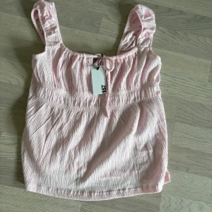 Ljusrosa blus med knyt från Angela - Supersöt ljusrosa blus från Angela i storlek M. Blusen har breda axelband med rynkade detaljer och ett litet knyt framtill vid halsen. Materialet är mjukt och krinklat, vilket ger en chill och trendig vibe. Perfekt för varma dagar och enkel att styla med jeans eller kjol.