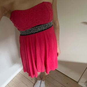 Bandeu Rosa klänning - Säljer en axelbandslös rosa klänning med spets upptill och ett glittrigt svart midjeband. Köpte på en secondhand butik utomlands och vet ej märket