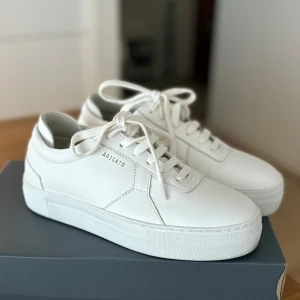 Vita sneakers från Axel Arigato - Snygga vita sneakers från Axel Arigato med stilren design och platt sula. Skorna har rund tå, snörning och är tillverkade i slätt skinn. Perfekta för dig som gillar minimalistisk och clean look. Axel Arigato-logga på sidan och innersulan. Tyvärr är dessa skor alldeles för små för mig!