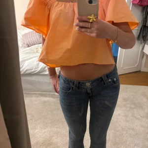 Orange croppad blus med volangärm - Superfin croppad blus i en härlig orange färg med vida volangärmar och offshoulder-modell. Toppen har en luftig och boxig passform som ger en riktigt somrig vibe. Perfekt för dig som vill sticka ut med färg och stil!
