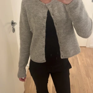 Grå stickad kofta från Gina Tricot - Mysig grå stickad kofta från Gina Tricot med rund hals och knappar hela vägen framtill. Koftan har lång ärm och är i en mjuk, lite fluffig kvalitet som känns skön mot huden. Perfekt att slänga på sig när det är kyligt ute.