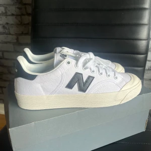 Vita New Balance sneakers med grå detaljer - Snygga vita New Balance sneakers med grått N på sidan och svart detalj vid hälen. Skorna har klassisk snörning och en platt, krämfärgad sula. Ovandelen är i slitstark canvas som ger en clean och fräsch look, perfekt för dig som gillar stilrena sneakers.