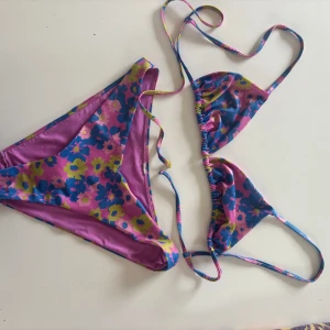 Blommig bikini i rosa och blått från WEEKDAY - Snygg bikini med blommigt mönster i starka färger som rosa, blått och gult. Toppen är trekantsmodell med smala snören att knyta i nacken och ryggen. Underdelen har klassisk form och samma färgglada blommor. Perfekt för stranden eller poolen i sommar!