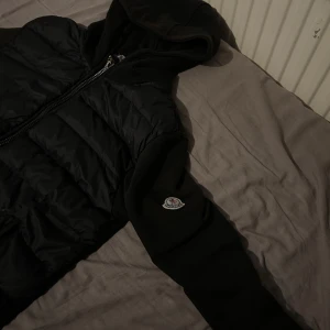 Svart pufferjacka från Moncler XL - Svart pufferjacka från Moncler i storlek XL. Jackan har huva, quiltad front och mjuka ärmar med Moncler-logga på vänster arm. Dragkedja framtill och stilren design som ger en clean vibe. Perfekt för dig som gillar exklusiva märken och streetwear.