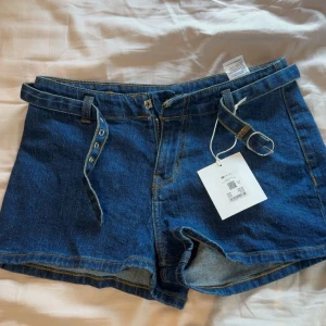 Blå jeansshorts med bälte från NA-KD - Snygga blå jeansshorts från NA-KD med tillhörande bälte i samma denimtyg. Shortsen har en rak passform och är perfekta för varma dagar. Tidlös design med kontrastsömmar och bälteshällor.