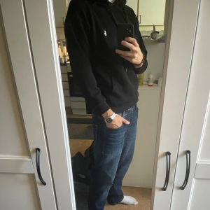 Svart hoodie från Polo Ralph Lauren - Svart hoodie från Polo Ralph Lauren med klassisk vit logobrodyr på bröstet. Tröjan har huva med snörning, magficka och långa ärmar. Mjuk bomullskvalitet och clean look som funkar till jeans eller joggers. Perfekt för chill dagar eller när du vill ha en enkel stil. Pris kan diskuteras 🍾