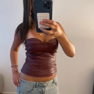 Vinröd korsett-topp i skinnimitation - Snygg vinröd korsett-topp med sweetheart-urringning och croppad passform. Gjord i skinnimitation som ger en cool och edgy vibe. Axelbandslös design som sitter tight och framhäver siluetten. Perfekt att styla med jeans eller kjol för en trendig look.