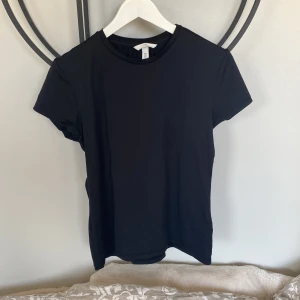 Svart t-shirt från H&M, storlek L - En stilren svart t-shirt från H&M i storlek L. Modellen har rund hals och korta ärmar, perfekt för en clean och enkel look. Materialet är mjukt och stretchigt, tillverkat av 96% polyamid och 4% elastan. Passformen är normal och t-shirten är helsvart utan mönster eller detaljer.